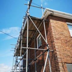 Metal scaffolding is erected alongside a brick house, reaching above the roofline under a blue sky with scattered clouds. This domestic scaffolding provides access for construction or maintenance work by a skilled scaffolder.
