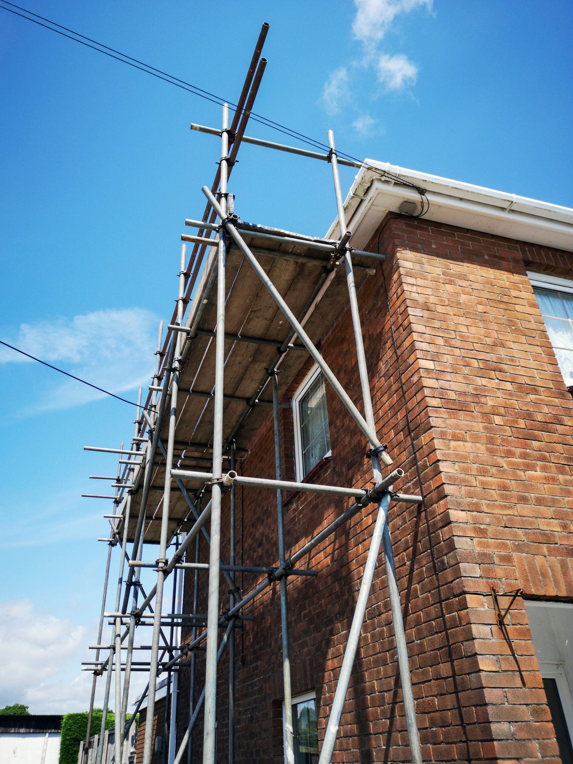 Metal scaffolding is erected alongside a brick house, reaching above the roofline under a blue sky with scattered clouds. This domestic scaffolding provides access for construction or maintenance work by a skilled scaffolder.
