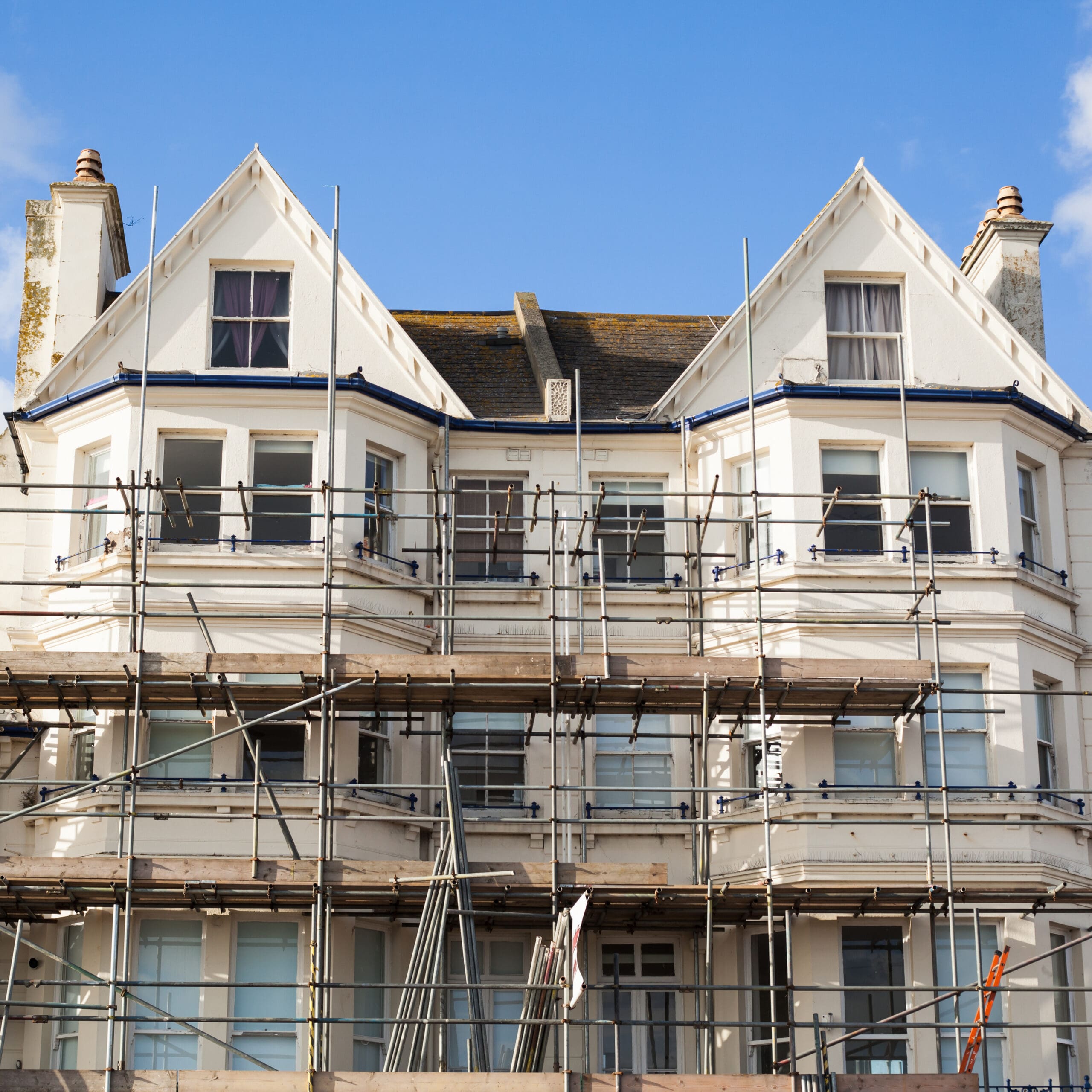 A large white house with multiple chimneys and gabled roofs is covered in domestic scaffolding, indicating ongoing renovation work by a skilled scaffolder, under a clear blue sky.