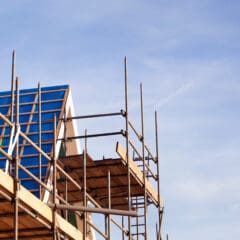 Domestic scaffolding surrounds a building under construction with a blue roof, set against a clear, blue sky.