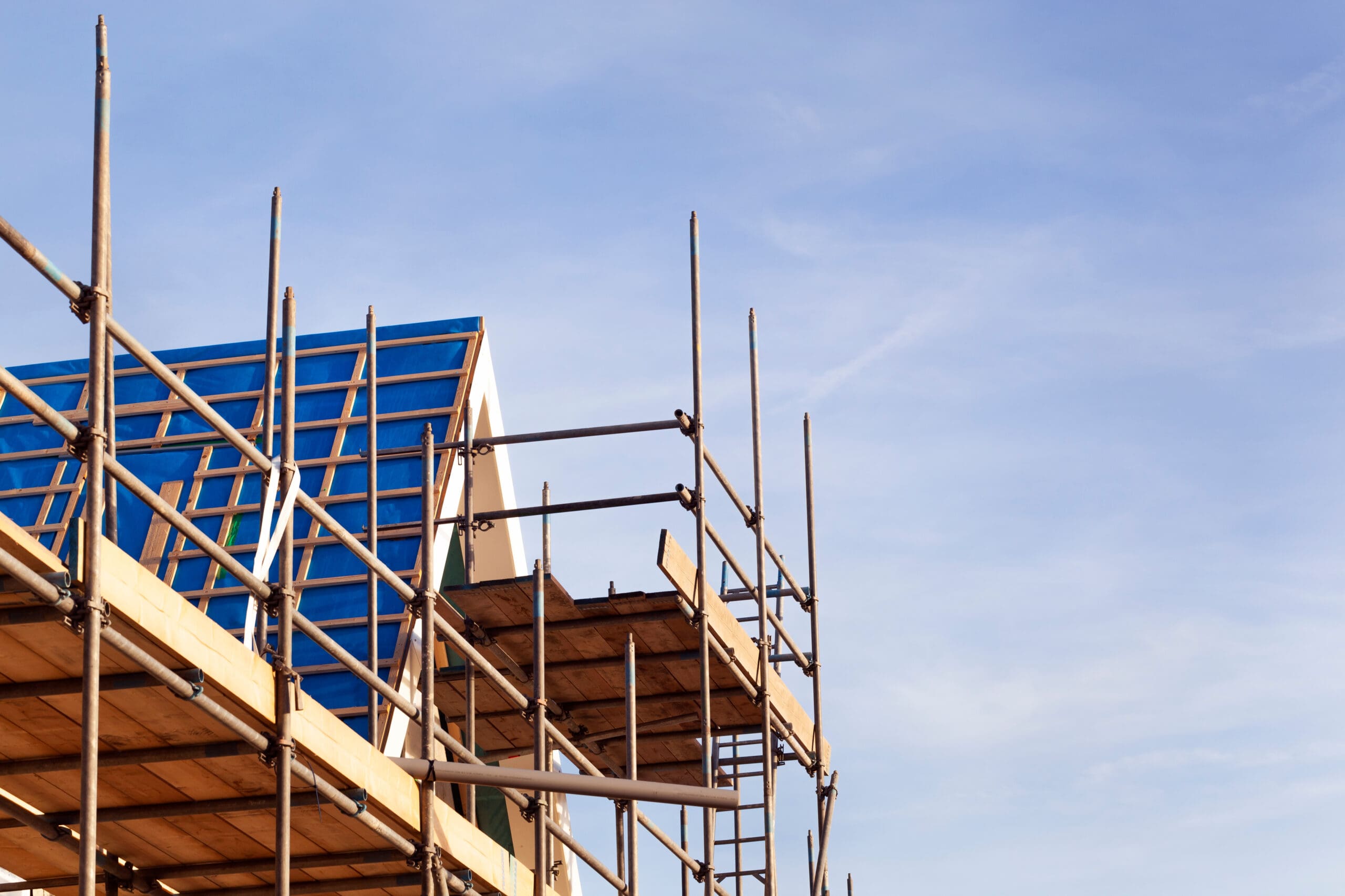 Domestic scaffolding surrounds a building under construction with a blue roof, set against a clear, blue sky.