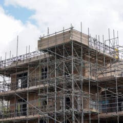 A multi-story building under construction, covered in commercial scaffolding, with brick walls visible. The sky above is partly cloudy, and green trees are seen on the left side of the image.