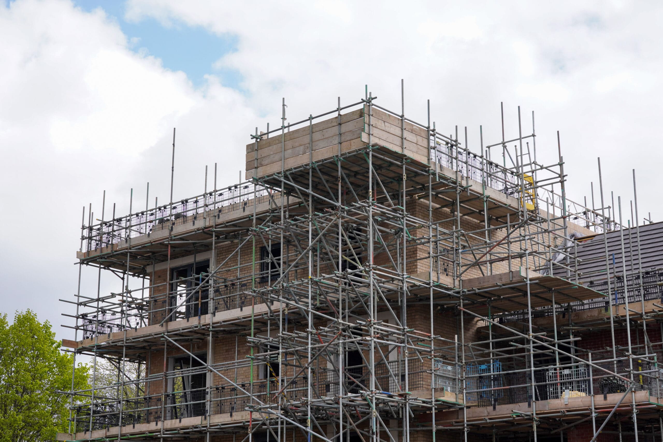A multi-story building under construction, covered in commercial scaffolding, with brick walls visible. The sky above is partly cloudy, and green trees are seen on the left side of the image.