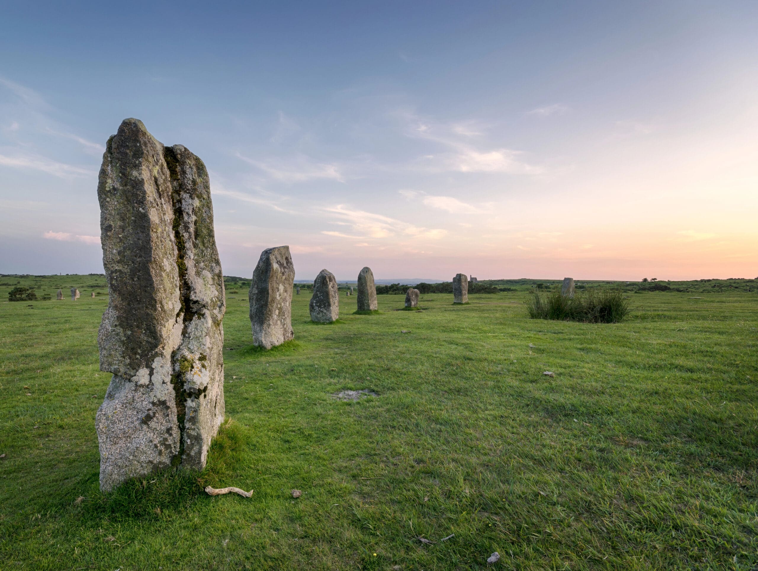 A row of large, ancient standing stones rises from grassy open land at sunset, their silhouettes as impressive as a well-constructed commercial scaffolding against the partly cloudy sky.